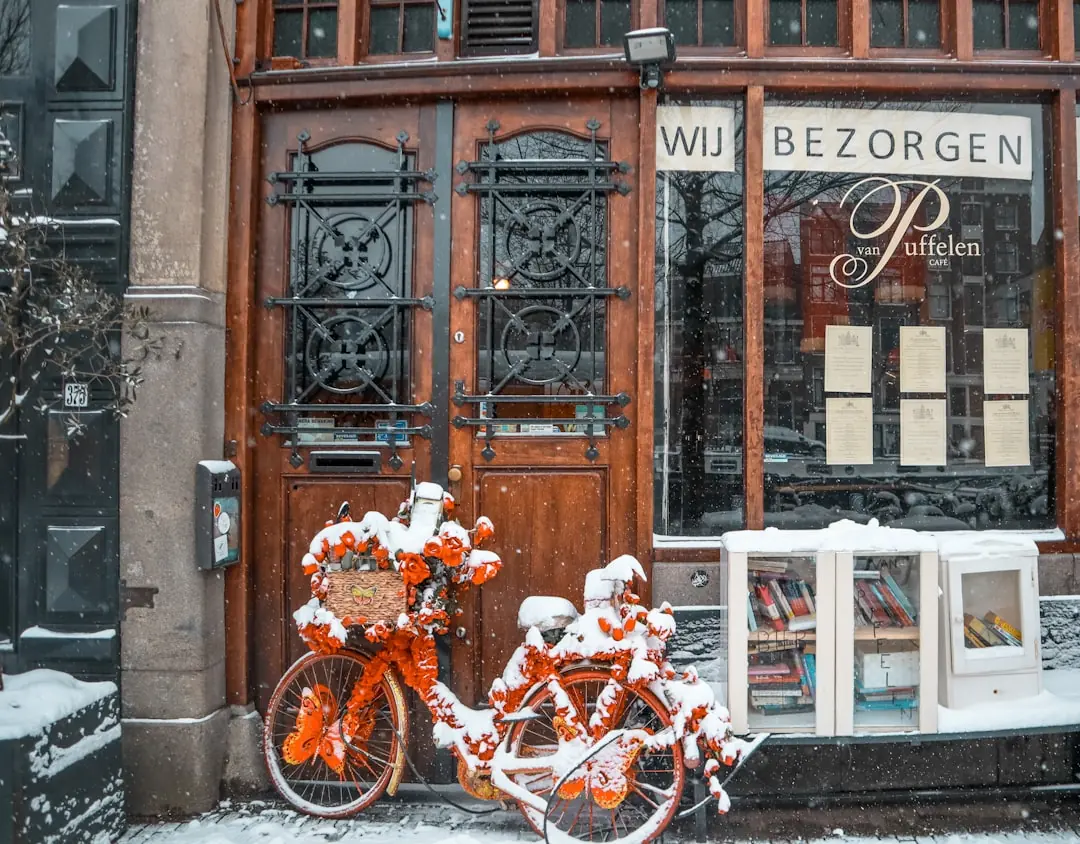 Schneebedecktes, mit orangefarbenen Blumen verziertes Fahrrad vor dem Café van Puffelen in Amsterdam bei Schneefall