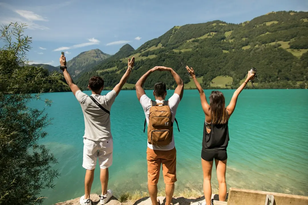 Drei junge Menschen stehen mit erhobenen Armen an einem türkisfarbenen Bergsee vor grüner Alpenkulisse