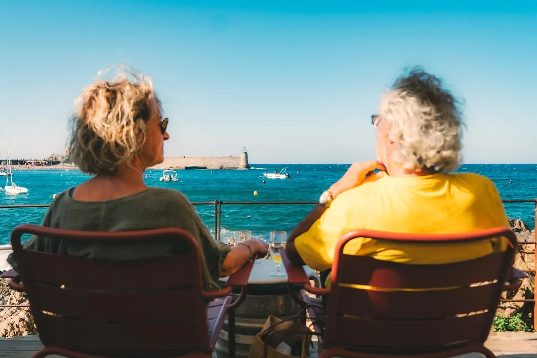 Zwei entspannte Personen sitzen in roten Stühlen und blicken entspannt von einer Terrasse auf das azurblaue Meer am Hafen