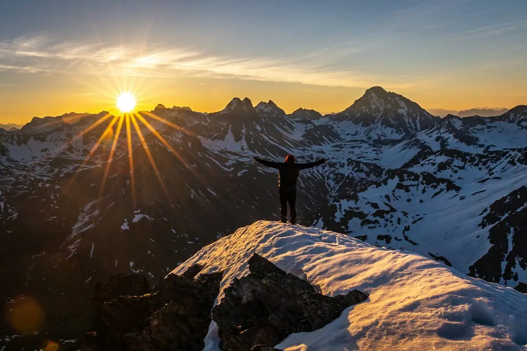Silhouette eines Wanderers mit ausgebreiteten Armen auf einem verschneiten Gipfel bei Sonnenaufgang in den Alpen