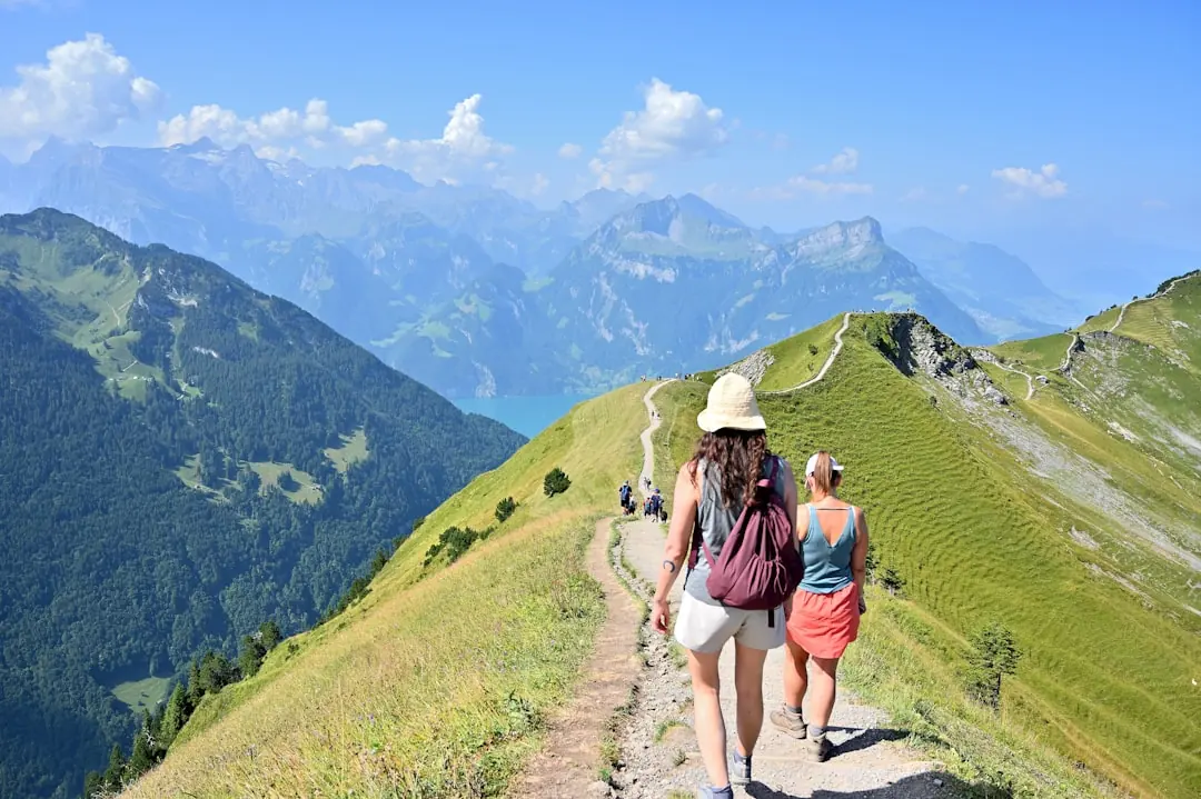 Zwei Frauen wandern auf einem schmalen Gratweg in den grünen Schweizer Alpen mit Blick auf einen tiefblauen Bergsee