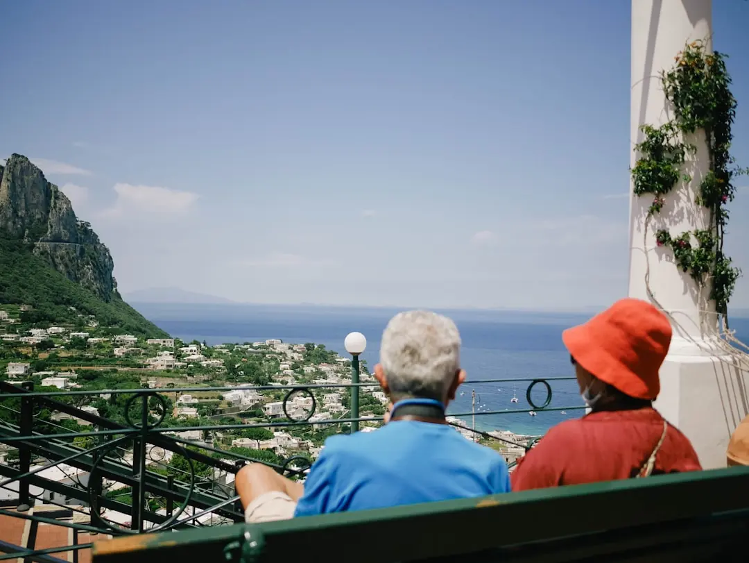 Ein Paar genießt von einer Terrasse den weiten Ausblick auf eine mediterrane Küstenstadt am tiefblauen Meer