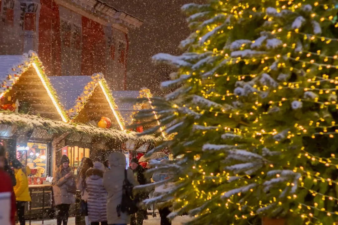 Verschneiter Weihnachtsmarkt bei Nacht mit beleuchteten Holzbuden, Besuchern und einem großen Christbaum im Vordergrund