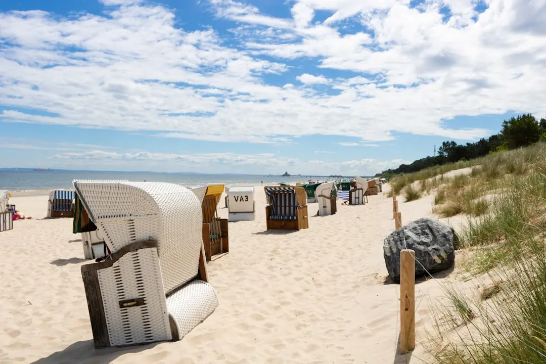 Weiße Strandkörbe an einem sonnigen Sandstrand der Ostsee vor bewachsenen Dünen unter blauem Himmel mit Schönwetterwolken